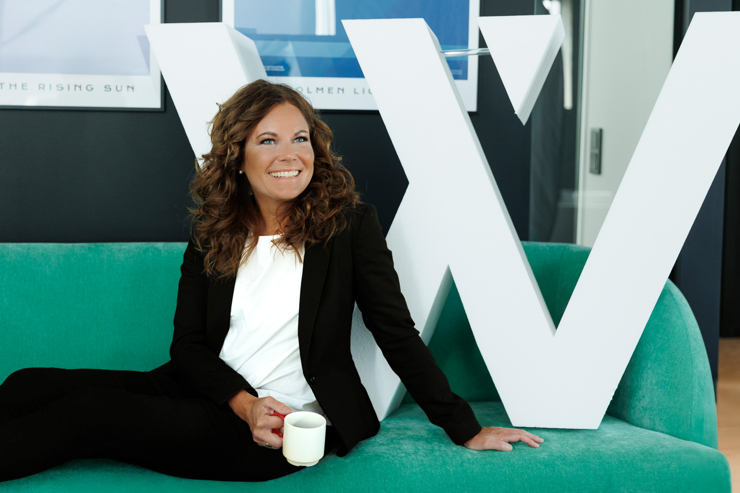 Annette Söby Hermansson sitter leende på en grön soffa med en kopp kaffe i handen, framför en stor vit bokstav "W" på Way Communications kontor.