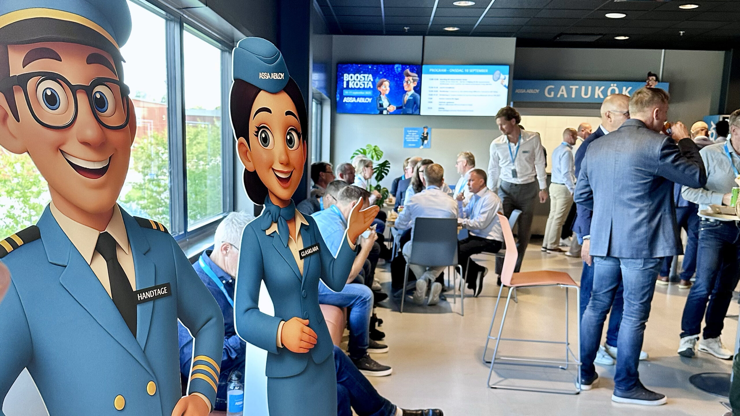Besökare minglar i en lounge under ASSA ABLOYs ‘Boosta i Kosta’; två blå uniformerade kartongfigurer i förgrunden och programskärmar i bakgrunden.